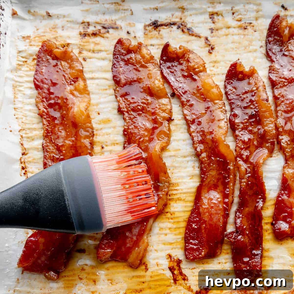 Maple Bacon Recipe with Just 2 Ingredients 7 A brush delicately applies a rich maple glaze to perfectly cooked bacon on a baking sheet, infusing each strip with sweet, smoky goodness.