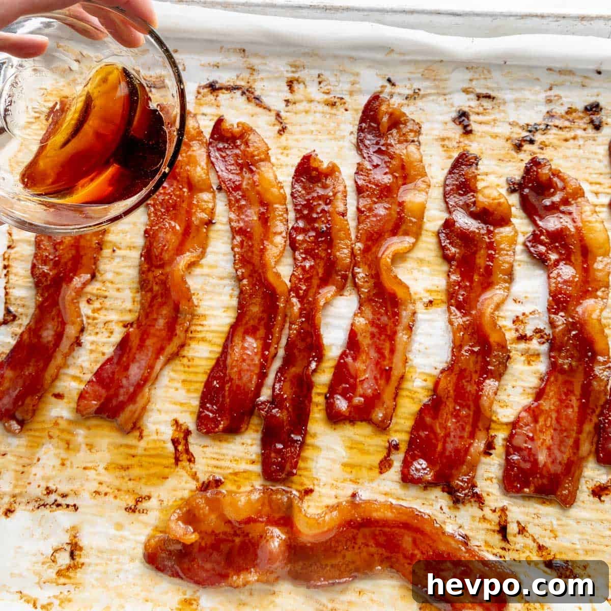 Maple Bacon Recipe with Just 2 Ingredients 8 Maple bacon strips on a baking sheet are being glazed with a rich sauce poured from a glass bowl.