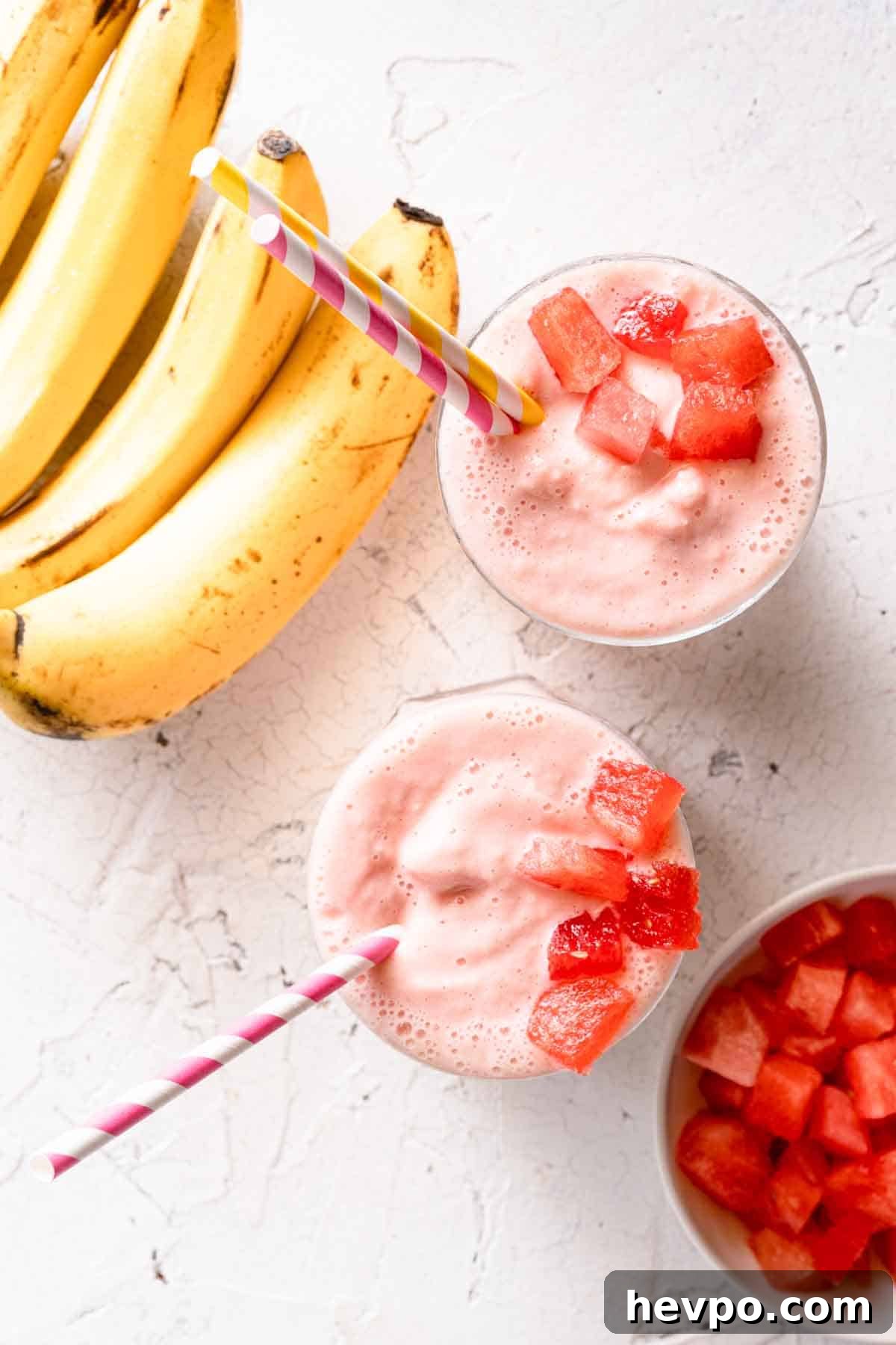 two smoothies garnished with chunks of watermelon with bananas and bowl of watermelon on the side
