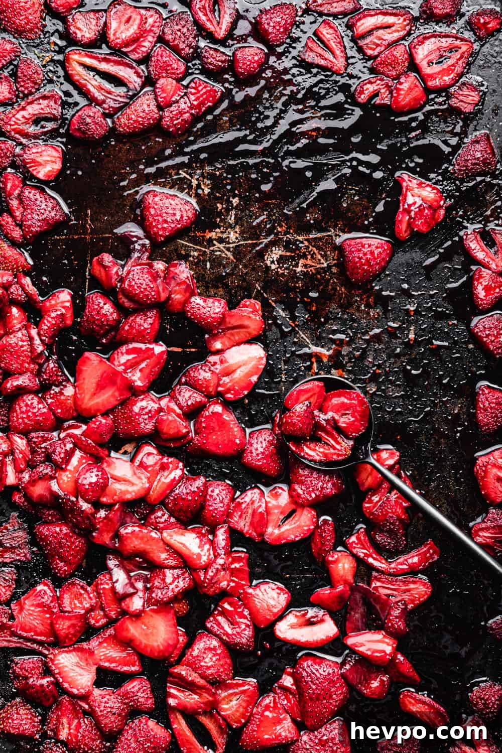 Roasted Strawberries: How to Cook and Use Them for Bold Flavor 13 Overhead shot of jammy roasted strawberries in their syrup on a dark sheet pan with a black spoon, post-oven, on a dark background.