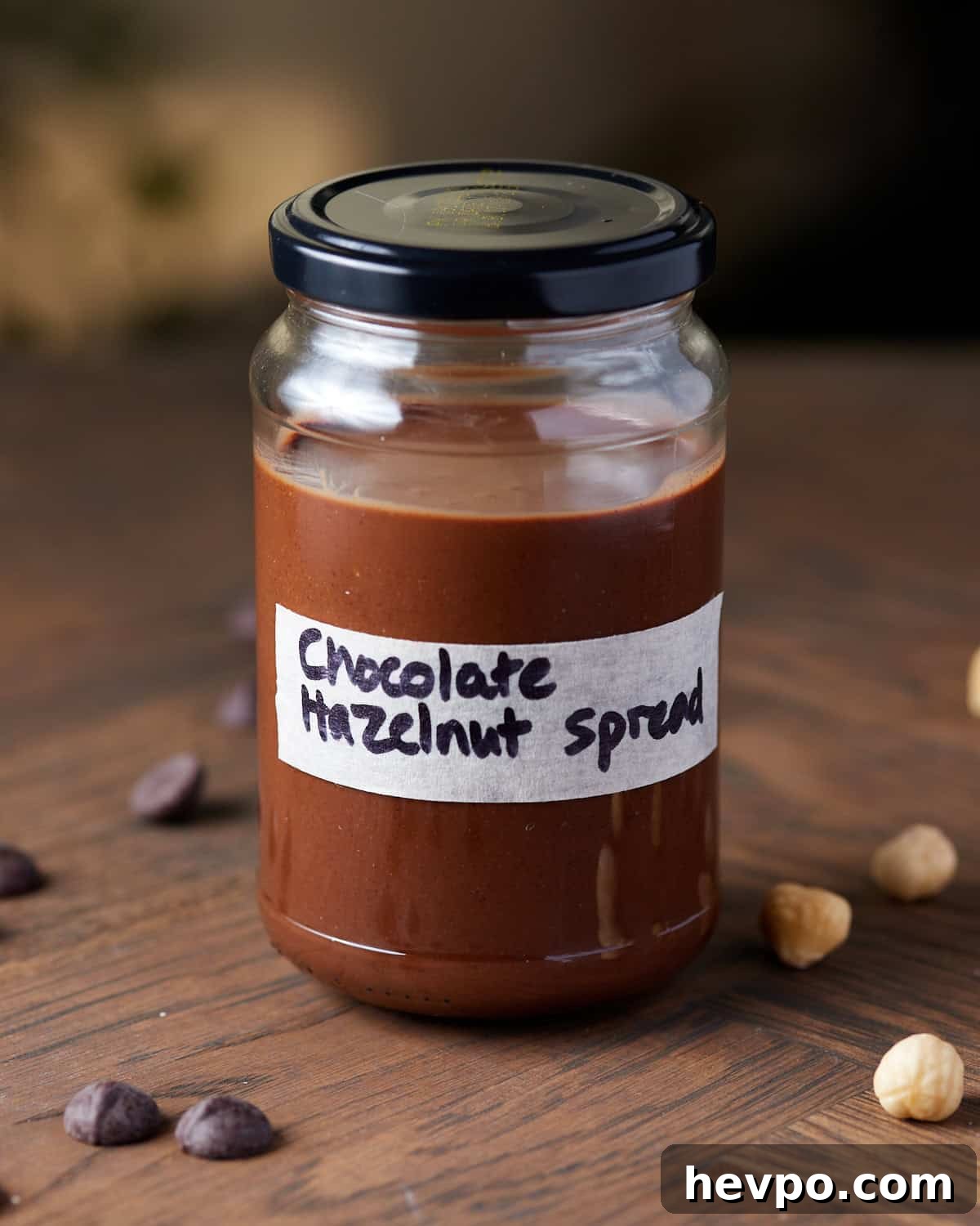 Photo showing a jar of homemade chocolate hazelnut spread with a hand-written label, set on a wooden surface with hazelnuts and chocolate chips scattered around.
